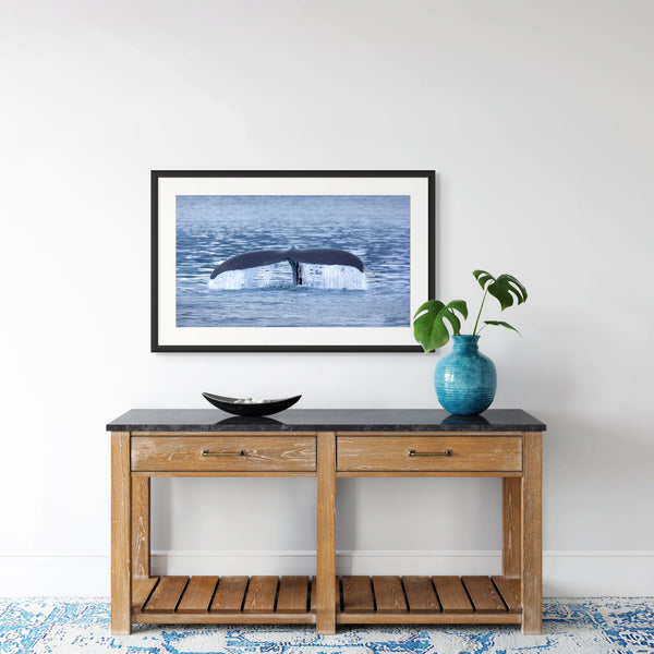 Framed whale tail artwork on a wall above a wooden console table with decorative items.