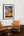 Framed artwork of a sunset with two wooden chairs and a small table in the foreground.
