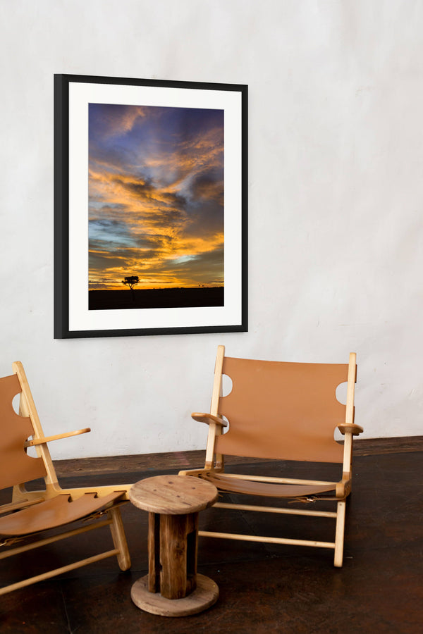 Framed artwork of a sunset with two wooden chairs and a small table in the foreground.