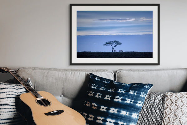 Framed landscape print of a tree on a savanna above a couch with a guitar and decorative pillows.