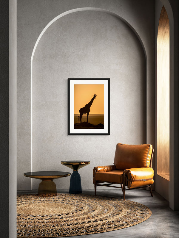 Framed artwork of a giraffe on a wall in a room with a brown leather chair and round table.