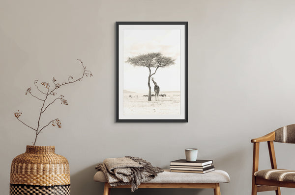 Framed artwork of a giraffe and tree on a wall above a bench with a vase and books.